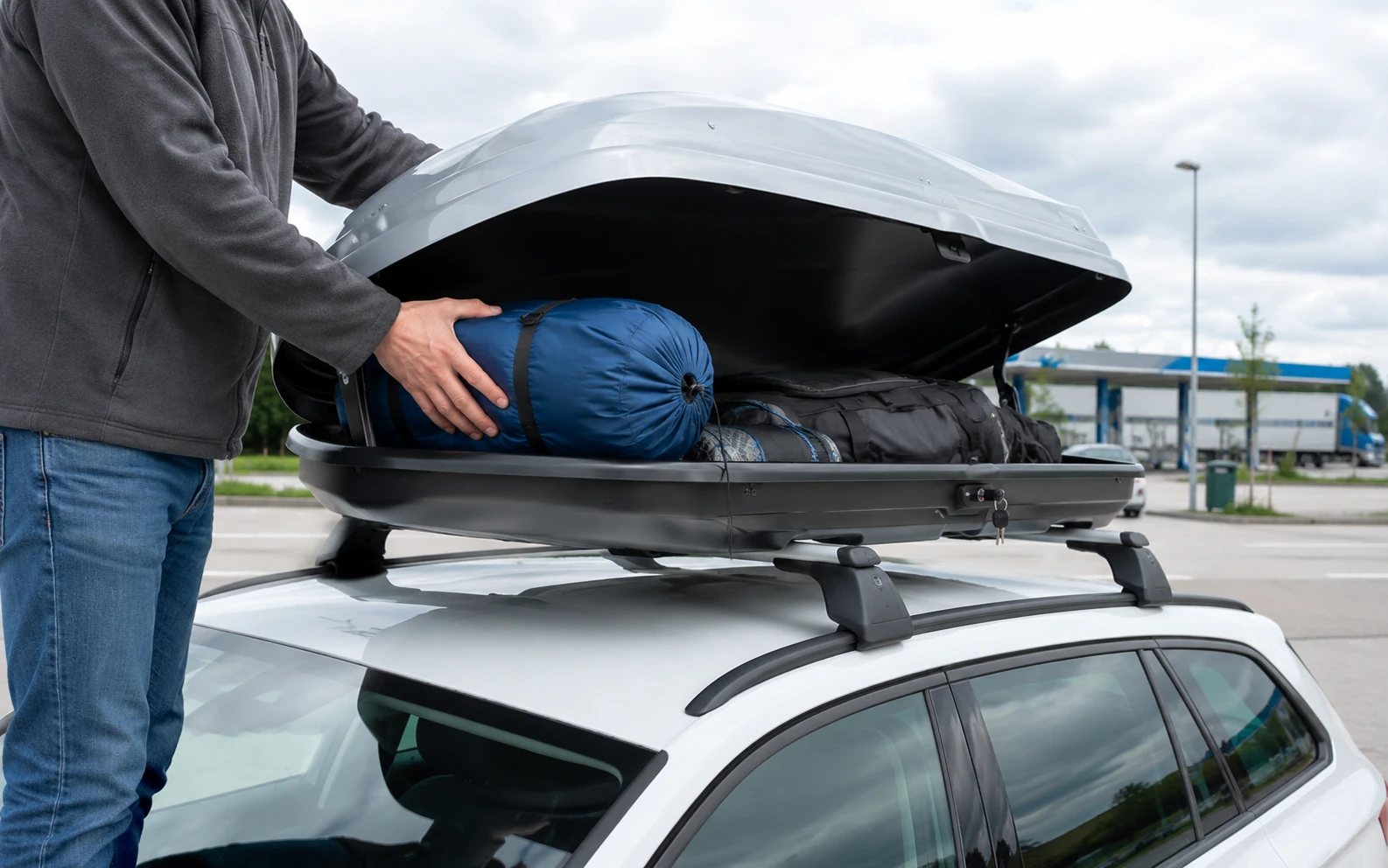 Eine Person packt leichte Schlafsäcke in eine silberne Dachbox auf einem weißen Kombi bei einer Autobahn-Raststätte