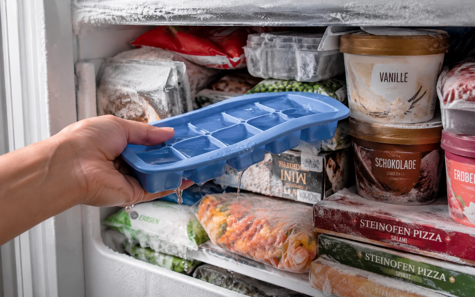 Eine Hand versucht frustriert eine vereiste Silikon-Eiswürfelform in ein überfülltes Gefrierfach zu quetschen