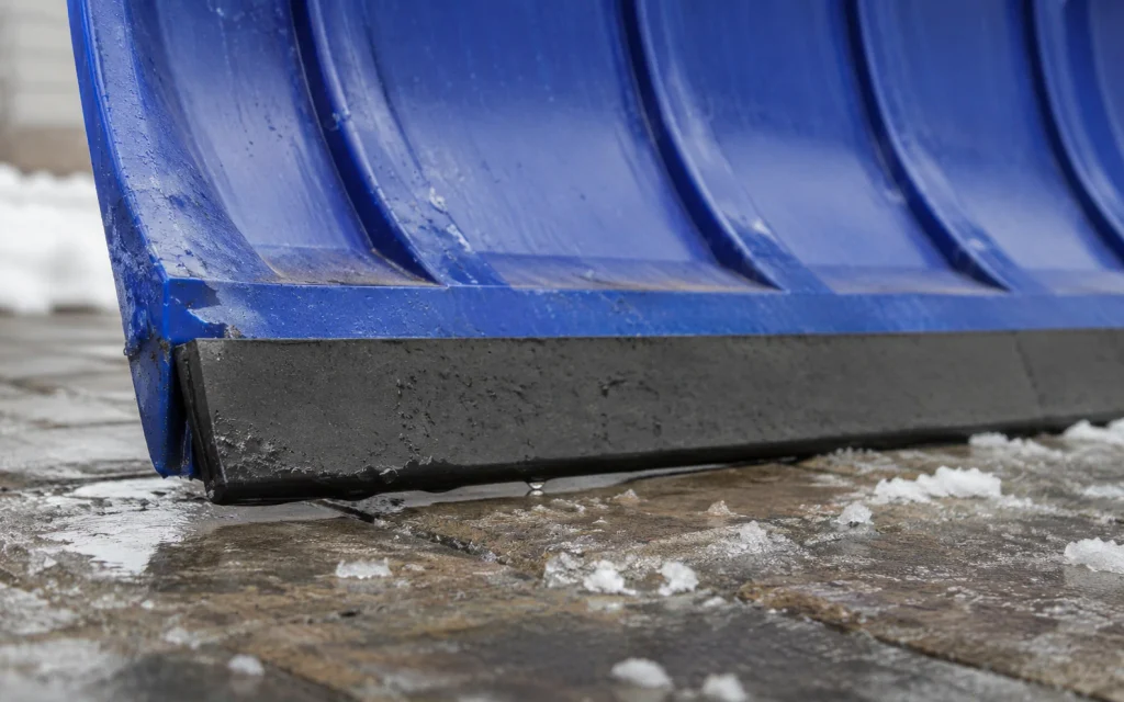 Nahaufnahme der Gummi-Gleitkante an der Schaufel eines Schneeschiebers auf gepflastertem Untergrund