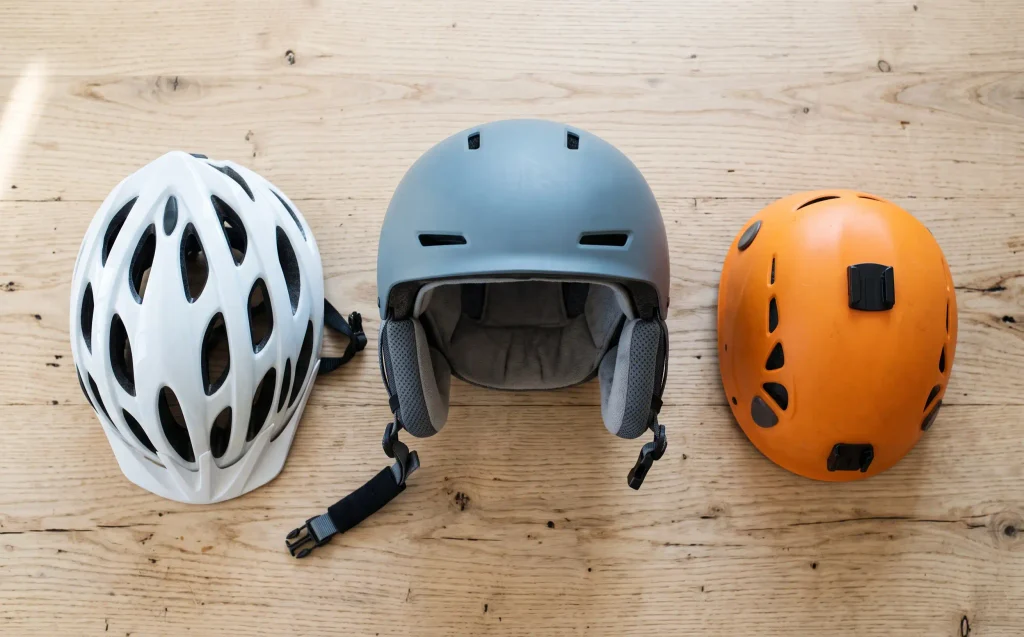 Skihelm, Fahrradhelm und Kletterhelm nebeneinander auf einem Holztisch zum Vergleich der Bauformen